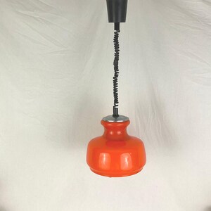 Puede incluir: Lámpara colgante de cristal naranja con cable y montaje en techo negros. La lámpara tiene una forma redondeada y bulbosa y un anillo metálico plateado en la parte superior. El cable es de diseño negro y enrollado.