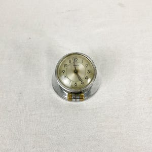 May include: A silver and gold-toned Omega clock with a white face and black hands. The clock is designed to be attached to a surface.