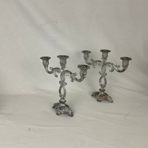 May include: Two silver-toned metal candle holders with ornate designs. Each holder has three candle arms and a base with a decorative floral pattern.