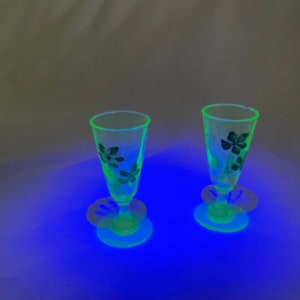 May include: Two clear, stemmed glasses with a floral design. The glasses have a flared shape and sit on a round base with a decorative, openwork design. The floral pattern is dark, and the glass glows green under a black light.