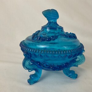 May include: A blue glass candy dish with a seashell design. The dish has three legs shaped like seahorses and a lid with a seashell finial.