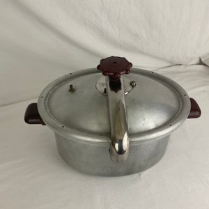 May include: A vintage oval-shaped aluminum pressure cooker with a red knob and a silver metal steam release valve. The cooker has two black handles.