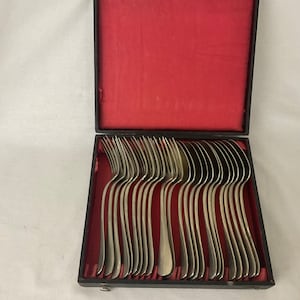 Può includere: Un set di forchette d'argento disposte ordinatamente in una scatola nera con rivestimento in velluto rosso. Le forchette hanno un design classico, con manici e rebbi curvi. La scatola è aperta, rivelando le posate.