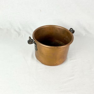 May include: A copper pot with two black metal handles. The pot is round and has a smooth surface.