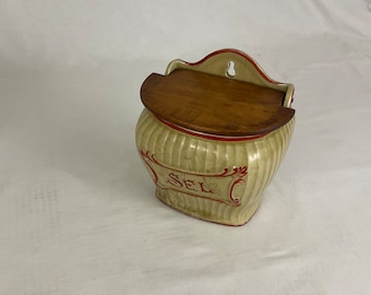 May include: Cream-colored ceramic salt cellar with a wooden lid. The salt cellar has vertical ribbed detailing and a red trim. The word "SEL" is written in red script on the front. A small hole is at the top for hanging.