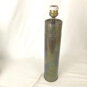 Puede incluir: Una lámpara de mesa cilíndrica con base color latón y casquillo blanco. La lámpara tiene un diseño decorativo en el lateral y un cable de alimentación. La lámpara mide aproximadamente 46 cm de altura.