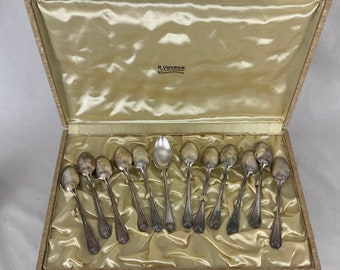 May include: A collection of silver spoons presented in a beige satin-lined box. The spoons are arranged in an orderly fashion, with different handle patterns. The box is open, displaying the spoons and the interior lining. The box has a clasp and the text "R. Veyron" is visible.