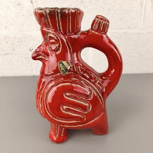 May include: A red ceramic pitcher in the shape of a rooster. The pitcher has a handle, a spout, and is decorated with embossed details. A small emblem is affixed to the front. The rooster-shaped pitcher is a decorative item.