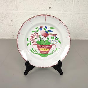 Könnte beinhalten: Weißer Keramikteller mit handgemaltem Vogel- und Blumendesign. Der Teller hat einen roten Rand und einen dekorativen Vogel über einem rot-gelben, becherförmigen Pflanzgefäß. Der Teller wird auf einem schwarzen Ständer präsentiert.