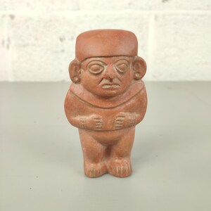 May include: A terracotta-colored figurine of a standing figure with a hat, large ears, and hands clasped at the chest. The figure has a stern expression and is likely a representation of a historical or cultural artifact.