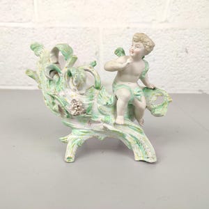 Może przedstawiać: Porcelanowa figurka cherubina siedzącego na dekoracyjnej, przypominającej drzewo strukturze. Cherubin jest nagi o jasnej karnacji, a struktura pomalowana jest w odcieniach zieleni i bieli. Figurka ma estetykę vintage.