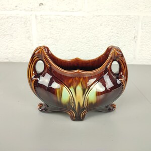 Può includere: Un vaso da fiori in ceramica smaltata marrone e verde con una forma unica e tre gambe. Il vaso ha due maniglie sui lati.
