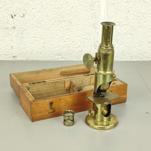 May include: A vintage brass microscope with a wooden storage box. The microscope has a cylindrical body with adjustable knobs and a circular base. A small brass eyepiece and a lens are also present. The wooden box is open, revealing its interior.
