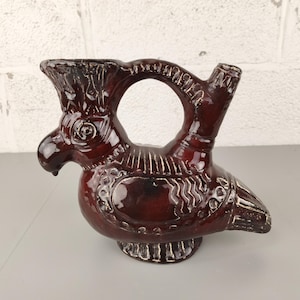 May include: A handcrafted, bird-shaped ceramic pitcher with a rich, dark red glaze. The pitcher features a detailed design with white accents, including a circular handle and a spout. The base is a pedestal design. The pitcher is approximately 8 inches tall.