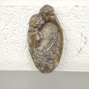 Op de afbeelding: Bronzen reliëfsculptuur met twee figuren in een omhelzing. De figuren hebben vloeiend haar en gedrapeerde stof, tegen een getextureerde achtergrond. De sculptuur heeft een ovale vorm.