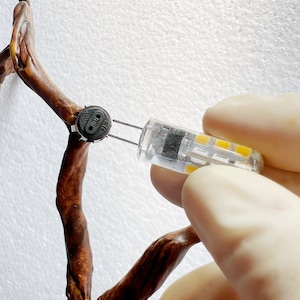 Puede incluir: Una bombilla LED transparente con filamentos amarillos se sostiene cerca de una rama marrón y texturizada. La bombilla tiene dos clavijas metálicas. La rama es parte de una estructura más grande y de aspecto natural.