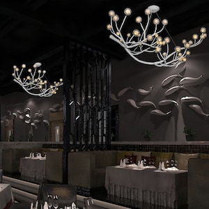 May include: Two white branch-shaped chandeliers with multiple round glass bulbs hang from the ceiling of a restaurant. The chandeliers are lit and cast a warm glow on the tables and chairs below.