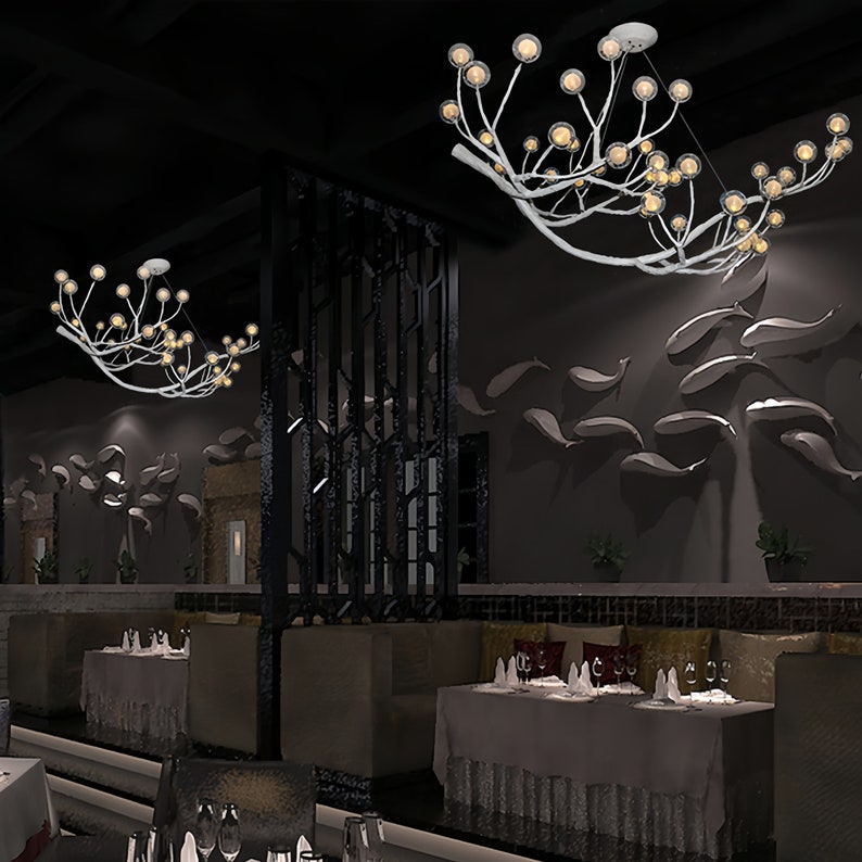 May include: Two white branch-shaped chandeliers with multiple round glass bulbs hang from the ceiling of a restaurant. The chandeliers are lit and cast a warm glow on the tables and chairs below.