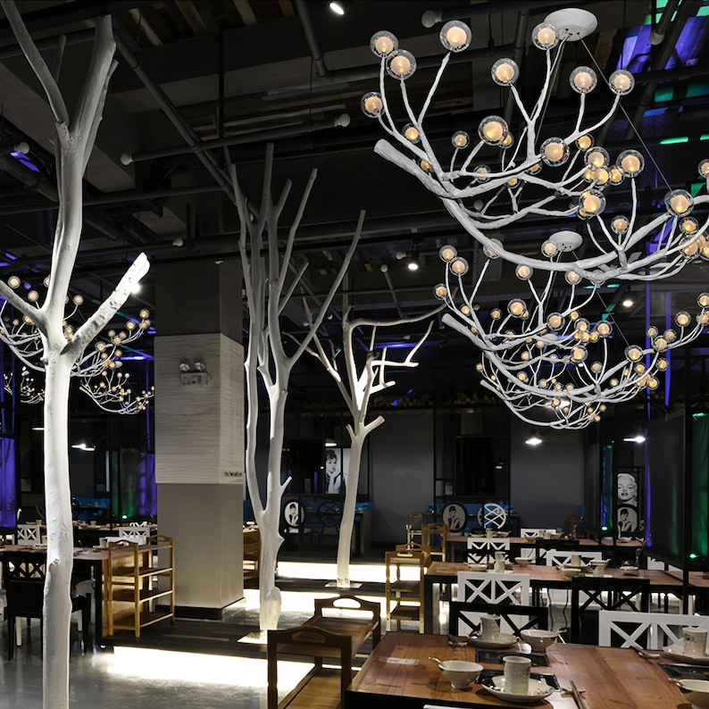May include: A modern restaurant interior with white faux trees and a unique white branch chandelier with multiple round light bulbs. The restaurant has wooden tables and chairs, and a dark grey tiled floor.