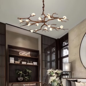 May include: A brown, branch-shaped chandelier with multiple white, flower-like light fixtures. The fixture hangs from a brown cord and is suspended from a white ceiling. The chandelier is in a room with dark wood furniture and a large window.