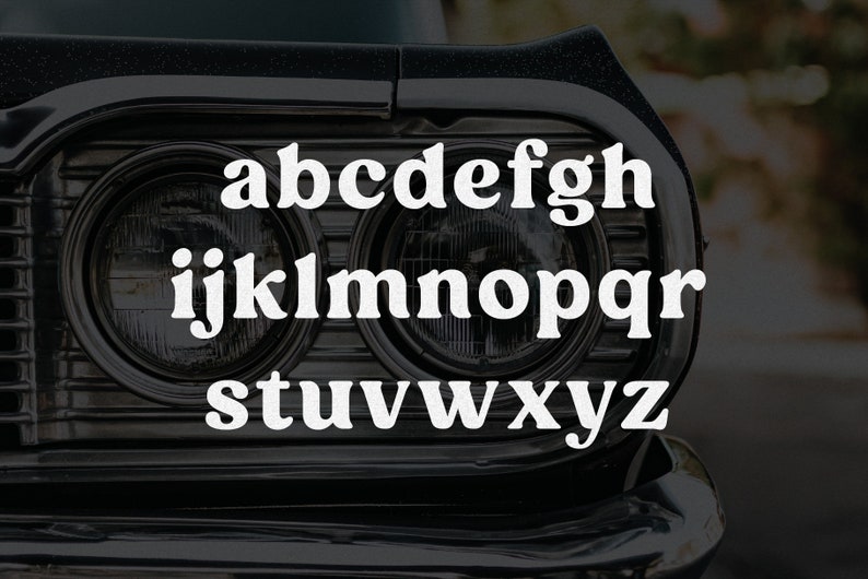 May include: A black car front grille with dual circular headlights featuring the lowercase alphabet letters a through z in a bold white serif font overlaid across the center of the vehicle's front end design.