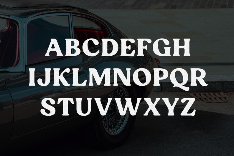 May include: A dark-colored vintage sports car with wire-spoke wheels is partially visible behind a white, bold, serif-style alphabet from A to Z arranged in three rows. The letters are clearly legible against the dark background of the car body.