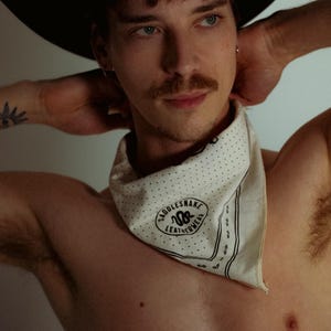 May include: A black cowboy hat with a silver band is worn. A white bandana with black polka dots and the text "Saddlesnake Leatherwear" is around the neck. The bandana has a black border. The image is a portrait.