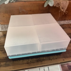 May include: A clear, square plastic container with four compartments. The container has a light blue trim and sits on a black base. The lid is frosted, and the container is on a wooden surface.