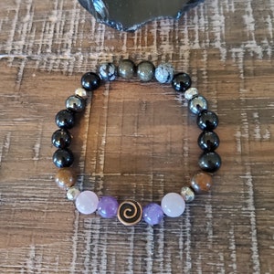 May include: A beaded bracelet with black, brown, grey, and purple beads. The bracelet has a silver charm with a spiral design.