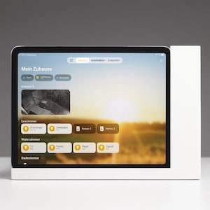 Könnte beinhalten: Ein Tablet, das eine Smart-Home-Steuerungsoberfläche anzeigt, mit den Worten "Mein Zuhause" oben. Der Bildschirm zeigt verschiedene Steuerungsoptionen und einen Live-Kamera-Feed. Das Tablet steht in einem weißen Ständer.