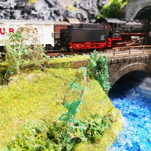 May include: A model train scene featuring a black and red steam locomotive pulling white freight cars with red text. The train crosses a stone bridge over a blue river, entering a tunnel. Green foliage and grass surround the tracks.