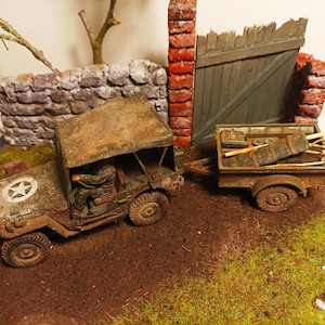 Può includere: Una jeep militare verde usurata con una capote in tela e una stella sul lato sta trainando un rimorchio carico di rifornimenti. La jeep è parcheggiata davanti a un muro di pietra e un cancello di legno.