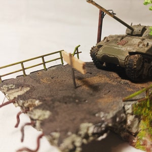 Puede incluir: Un modelo de tanque militar en un diorama con un poste indicador de madera, una valla verde y un pequeño árbol. El tanque está pintado con un patrón de camuflaje y tiene un cañón largo.
