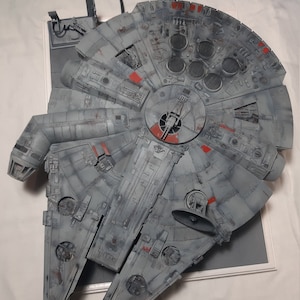 May include: A gray and silver model of the Millennium Falcon spaceship from Star Wars. The model is detailed with various panels and hatches, and has a weathered appearance.