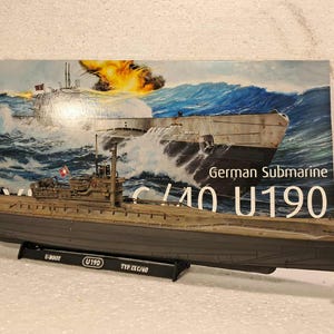 以下が含まれることがあります： ドイツのUボート潜水艦U190の模型。グレーの船体と甲板が特徴です。海戦シーンを描いた背景の前に展示されています。「German Submarine C/40 U190」の文字が見えます。