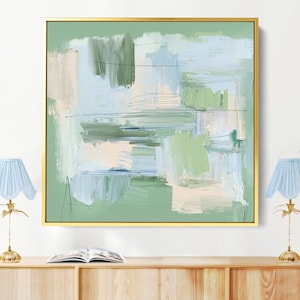 May include: Abstract artwork in a gold frame with green, blue, and beige brushstrokes. Two light blue lamps with gold bases sit on a light wood cabinet. A book is placed on the cabinet. The cabinet is made of light wood.