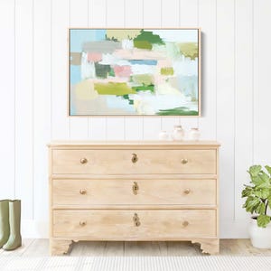 May include: A light wood chest of drawers with three drawers, brass knobs, and an abstract painting above. The painting has pastel colours. Green wellington boots stand on the floor near the chest.