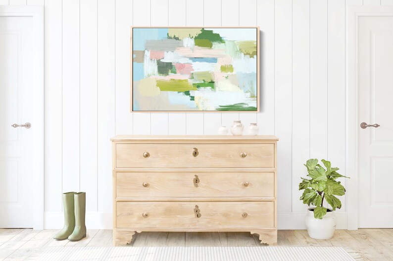 May include: A light wood chest of drawers with three drawers, brass knobs, and an abstract painting above. The painting has pastel colours. Green wellington boots stand on the floor near the chest.