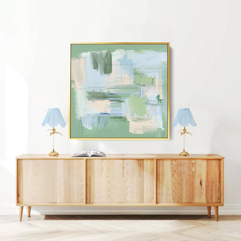 May include: Abstract artwork in a gold frame with green, blue, and beige brushstrokes. Two light blue lamps with gold bases sit on a light wood cabinet. A book is placed on the cabinet. The cabinet is made of light wood.
