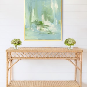 May include: An abstract painting in a gold frame, featuring shades of blue, green, and white. Below, a natural wicker console table with two shelves, and two small vases with green flowers. The wall is white.