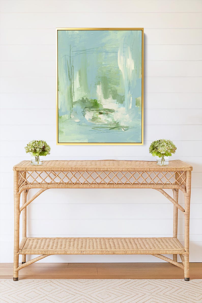 May include: An abstract painting in a gold frame, featuring shades of blue, green, and white. Below, a natural wicker console table with two shelves, and two small vases with green flowers. The wall is white.
