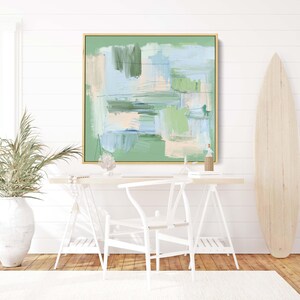 May include: Abstract art painting in a gold frame with shades of green, blue, and beige. The artwork hangs above a white desk with a white chair. A wooden surfboard leans against the wall.