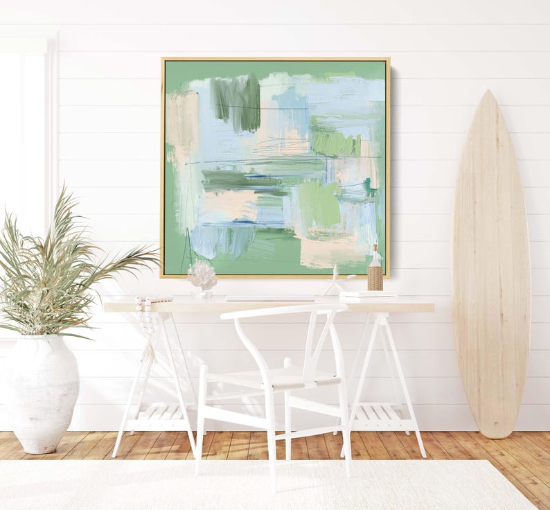 May include: Abstract art painting in a gold frame with shades of green, blue, and beige. The artwork hangs above a white desk with a white chair. A wooden surfboard leans against the wall.