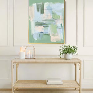 May include: Abstract art print in a gold frame, with green, blue, and beige brushstrokes. The artwork hangs above a light-coloured rattan console table with a lower shelf. Decorative candles and a small potted plant are also visible.