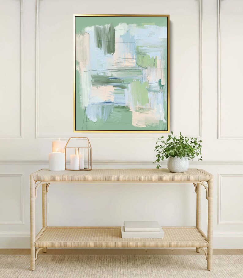 May include: Abstract art print in a gold frame, with green, blue, and beige brushstrokes. The artwork hangs above a light-coloured rattan console table with a lower shelf. Decorative candles and a small potted plant are also visible.