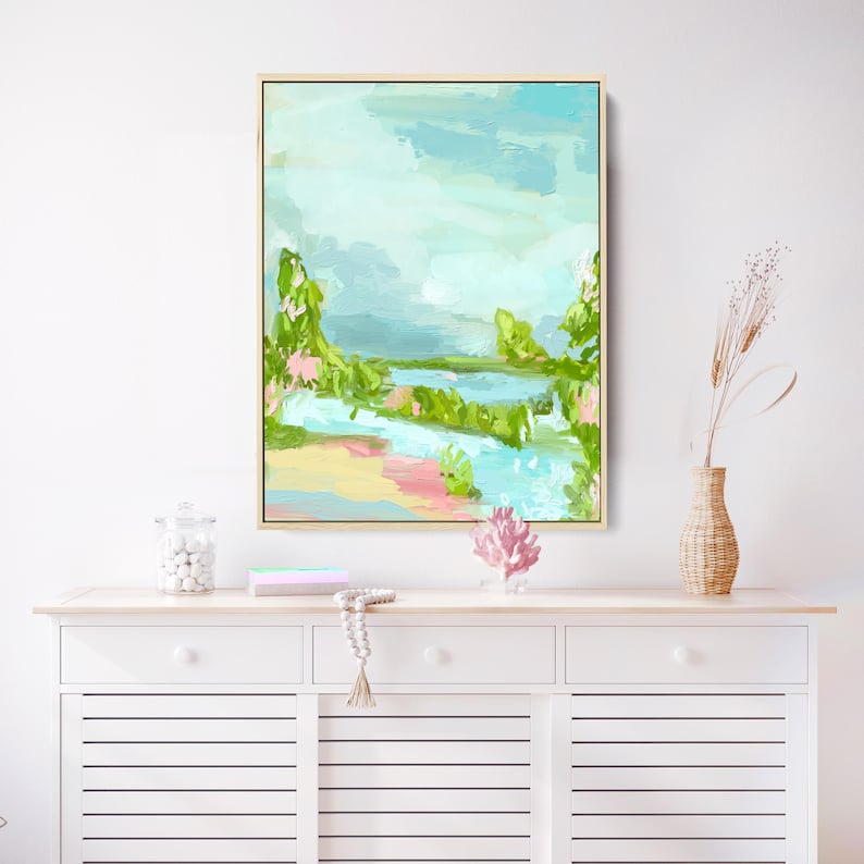 May include: Abstract landscape painting with a blue sky, green trees, and a pink and yellow beach. The painting is framed in a gold frame and is hanging on a white wall.