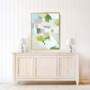 May include: Abstract art print in a gold frame. The artwork features brushstrokes of green, pink, blue, and grey on a light background. The painting hangs above a white cabinet with two blue and white lamps.
