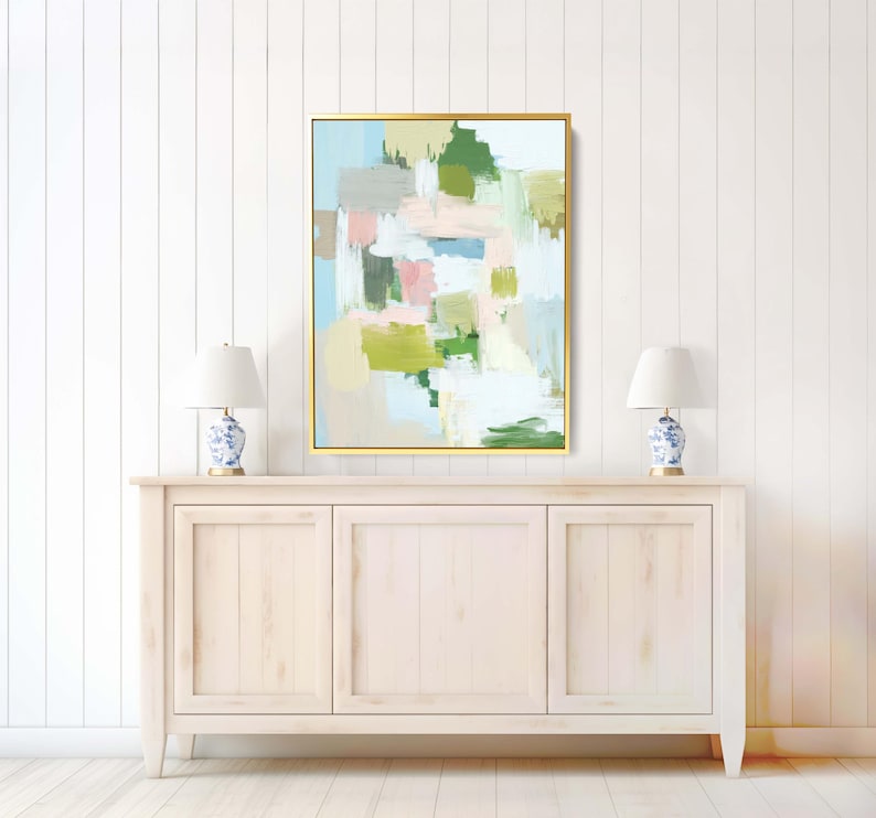 May include: Abstract art print in a gold frame. The artwork features brushstrokes of green, pink, blue, and grey on a light background. The painting hangs above a white cabinet with two blue and white lamps.
