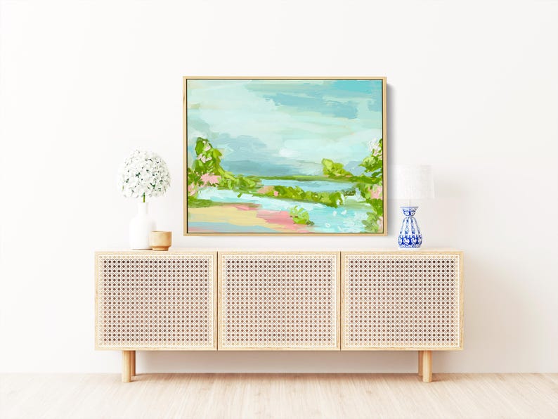 May include: A framed abstract landscape painting with a light blue sky, green trees, and a river. The painting is hanging on a white wall above a light wood cabinet with a woven rattan front.