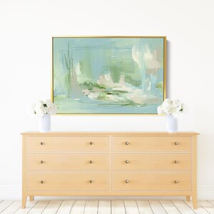 May include: An abstract painting in a gold frame hangs above a light wood dresser. The painting features soft blues, greens, and white brushstrokes. Two white vases with white flowers sit on the dresser, adding a touch of elegance.